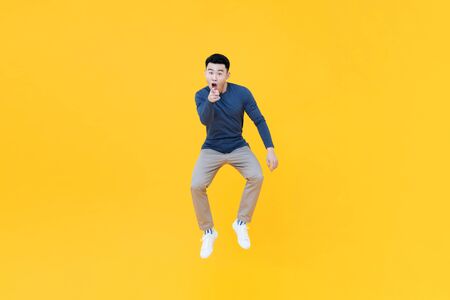 Full length portrait of young happy Asian man jumping in mid-air while in a fun pointing gesture on yellow isolated studio backgroundの写真素材