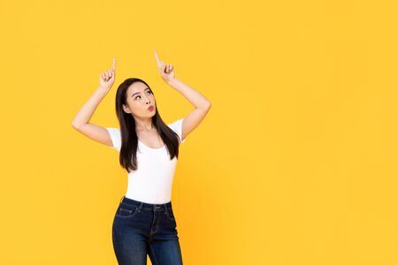 Young beautiful Asian woman pointing up with her two fingers while pouting her lips in a yellow isolated studio backgroundの写真素材