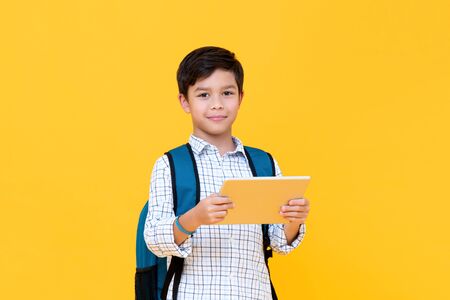Waist up portrait of handsome young schoolboy wearing backpack holding a tablet while looking straight in camera in yellow isolated studio backgroundの写真素材