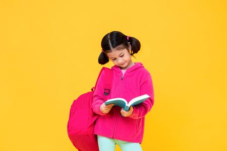 Portrait of cute pretty girl with pigtail hair in pink jacket and backpack reading book in yellow isolated studio backgroundの写真素材
