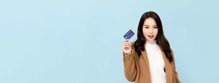 Beautiful smiling Asian woman in winter clothes showing credit card in hand for financial and cashless society concepts isolated on light blue banner backgroundの写真素材