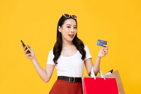 Beautiful smiling Asian woman with shopping bags and smart phone showing credit card in hand isolated on colorful yellow backgroundの写真素材