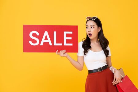 Shocked beautiful Asian woman carrying shopping bags showing red sale sign isolated on yellow backgroundの写真素材