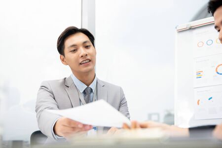 Cheerful Asian businessman suggesting business plan document to client at the meeting in the officeの写真素材