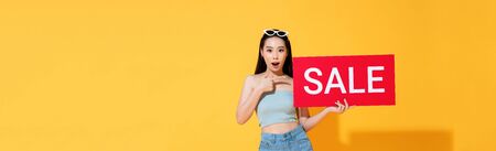 Shocked Asian woman in summer casual clothes pointing to red sale sign in hand isolated on yellow banner backgroundの写真素材