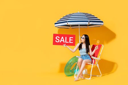 Surprised beautiful Asian woman in summer outfit holding red sale sign while sitting on beach chair isolated on yellow background with copy spaceの写真素材