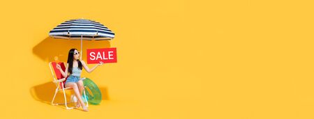 Surprised beautiful Asian woman in summer outfit holding red sale sign while sitting on beach chair isolated on yellow banner background with copy spaceの写真素材