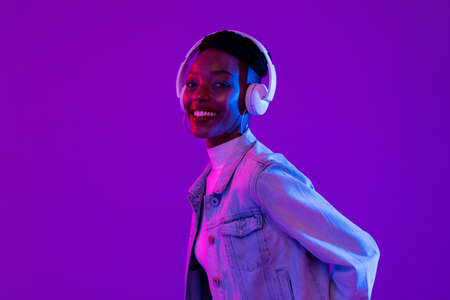 Stylish happy smiling African-American woman wearing headphones and listening to music in modern purple studio backgroundの写真素材