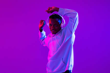 Young African man wearing wireless headphones listening to music and dancing in studio modern neon light purple backgroundの写真素材