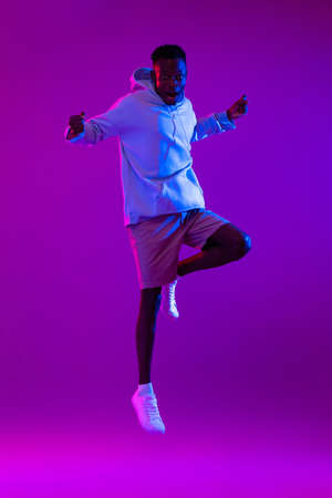 Full body portrait of ecstatic African man jumping in modern neon light studio backgroundの写真素材