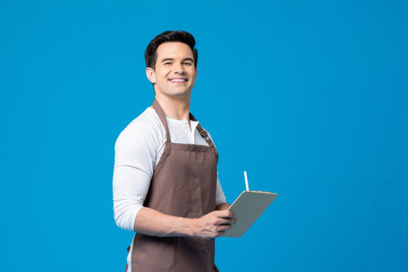 Cauca man with apron holding writing pad and pen in his hands while standing on blue background in light studioの写真素材