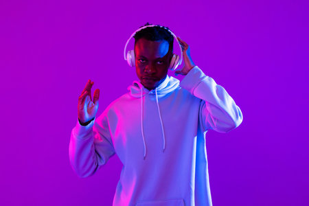 Young African man wearing wireless headphones listening to music in studio modern neon light purple backgroundの写真素材