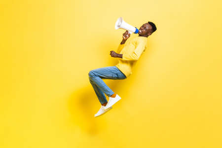 Full body side view of African angry man screaming in megaphone and looking at camera while levitating on yellow backgroundの写真素材