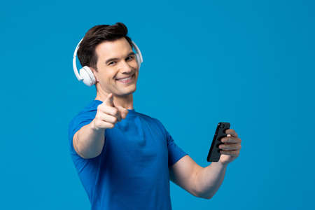 Smiling happy Caucasian man wearing headphones listening to streaming music on mobile phone in blue isolated studio backgroundの写真素材