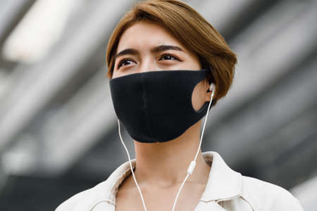 Young Asian woman wearing face mask outdoors while listening to music in the city during COVID-19 pandemicの写真素材