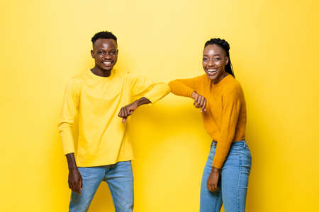 Happy young African American couple greeting with elbow bump in yellow color isolated studio backgroundの写真素材