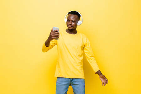 Happy young African man wearing headphones listening to music online from mobile phone in isolated yellow studio backgroundの写真素材