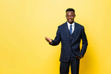 Positive African man in formal business suit smiling and opening hand doing welcome gesture in yellow color studio isolated backgroundの写真素材