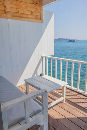 A view of the resort's balcony overlooking the sea, where the terrace features wooden sofas and white tables, and has wooden floors and ray of sunshine.の写真素材