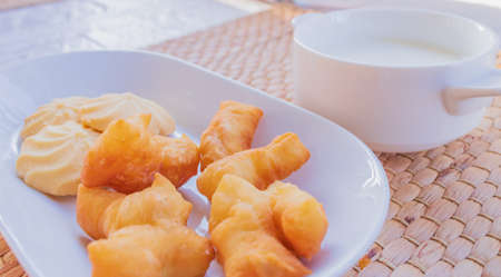 Close-up of breakfast on the dining table with deep-fried dough stick and cookies placed on a white plate with a glass of milk in the background.の写真素材