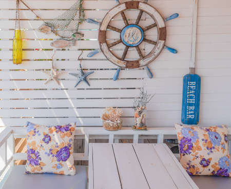 A picture of the exterior of the room, with a ship's steering wheel and starfish toys hanging on a white wooden wall and two pillows resting on a large white wooden sofa with a white tableの写真素材
