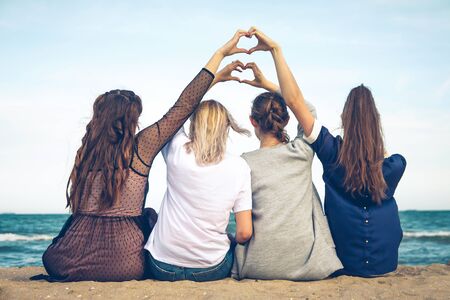 four girls sit on the seashore and show their hands a heart sign over their headsの写真素材