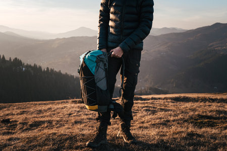 A hiker stands on a mountain ridge holding their backpack, ready to continue their journey. The golden light of the setting sun highlights the rugged terrain and the adventurer's determination.の写真素材