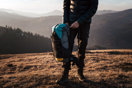 A hiker stands on a mountain ridge holding their backpack, ready to continue their journey. The golden light of the setting sun highlights the rugged terrain and the adventurer's determination.の写真素材