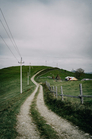 Rural dirt road leading to farm with haystacks and rolling hills under cloudy skyの写真素材