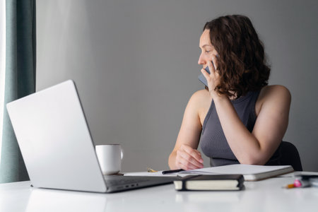 Woman managing work calls while sitting at home with laptop and notebookの写真素材