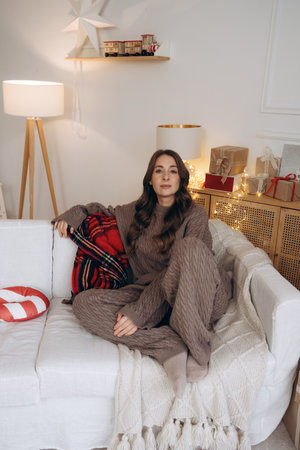 Cozy woman sitting on a sofa with Christmas gifts and decorations in the backgroundの写真素材