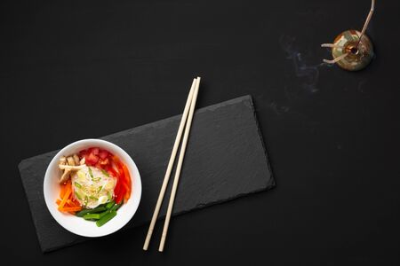 On a slate board in a white plate are glass funchoza noodles with fried chicken, red pepper, tomatoes, carrots, green beans and onions. Smoke from incense sticks. Black background. Top view Copy spaceの写真素材