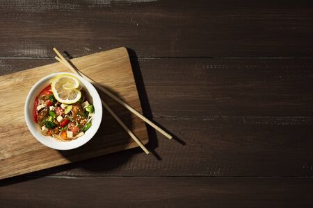 On a wooden brown background in a white plate are glass funchoza noodles with fried chicken, red pepper, tomatoes, carrots, lemon, green beans and onions. Moonlight. Rustic. Top view. Copy spaceの写真素材