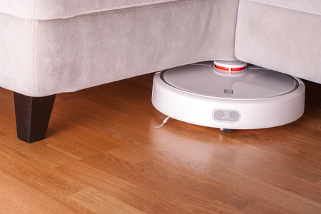 robotic vacuum cleaner runs under sofa in room on laminate floor modern smart cleaning technology housekeeping.の写真素材
