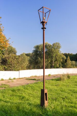 Old street lamp with broken light bulb and torn electrical wires on rusty metal pole in abandoned Park area against background of bright, blue sky, green grass and autumn forest.の写真素材
