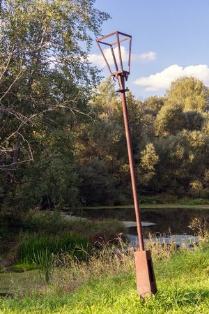 Old park. Against background of beautiful, dense green trees and dark surface of pond stands rusty, leaning, broken electric lantern. Summer landscape.の写真素材