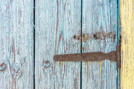Old rusty iron hinge with nails on wooden door with cracked peeling blue and yellow paint close up. Rural background.の写真素材