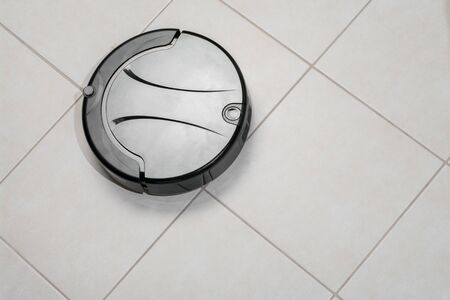 black robotic vacuum cleaner running on tile floor in kitchen. modern smart appliance for cleaning house.の写真素材