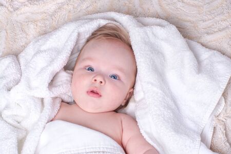 Surprised, newborn blue-eyed boy with plump lips and blond hair lies in dry, white towel on light fluffy blanket after bathing in bathroom. Textiles and bedding for children. Concept baby hygiene.の写真素材