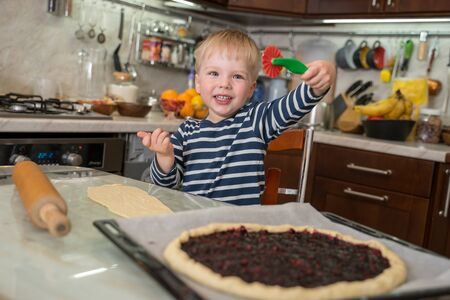 Little cute cheerful smiling blond boy holds in his hand children's shaped knife for cutting dough for making berry pie. Concept of happy childhood.の写真素材