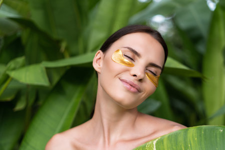 Close beauty portrait of a topless woman with perfect skin and natural make-up, shiny patches under the eyes, to moisturize the skin posing in tropical green leaves outdoorの写真素材