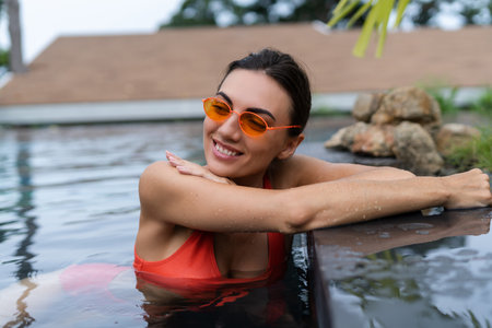 Beautiful european women in two pieces orange bikini swimsuit, tanned, with trendy sunglasses outside at tropical swimming poolの写真素材