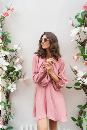 Young beautiful smiling cute romantic woman in trendy summer pink dress. Carefree woman posing in the street near white wall with flowers. Positive model outdoors in sunglasses.の写真素材