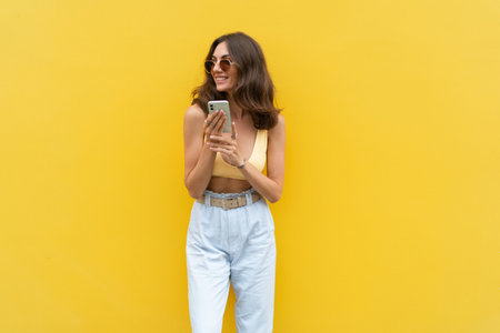 Young beautiful smiling woman with mobile phone. Carefree woman posing in the street near yellow wall. Positive model outdoors in sunglasses. Happy and excitedの写真素材