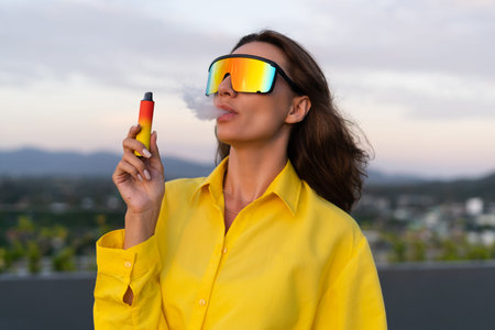 Stylish fit fashion women in bright yellow shirt trendy shield visor rainbow sunglasses posing at rooftop terrace tropical view sunset smoking vape pod enjoying puffsの写真素材