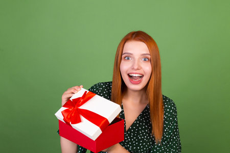 Young red hair woman on green background with gift box happy excited amazed surprisedの写真素材