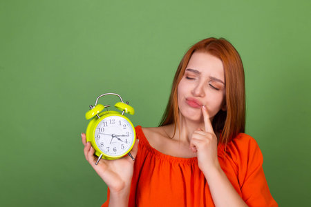 Young red hair woman in casual orange blouse on green background holding alarm clock thoughtful...