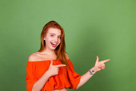 Young red hair woman in casual orange blouse on green background smile and laugh point rightの写真素材