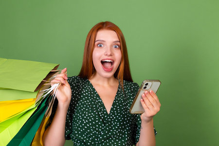 Young red hair woman on green background with shopping bags and mobile phone shocked amazedの写真素材