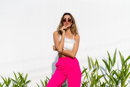 Stylish fit tanned beautiful woman in sunglasses, fashion pink pants and white top posing outdoor at white wall tropical leaves background, sunny day, natural lightの写真素材
